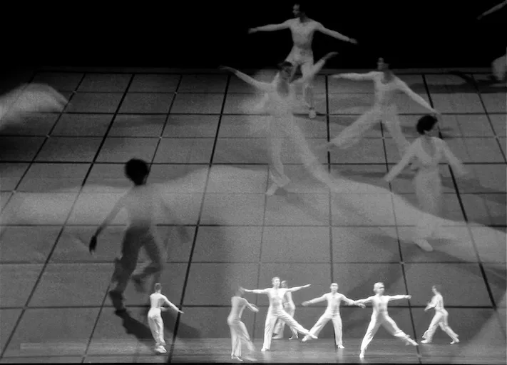 Black and white photograph of dancers in white moving across a gridded stage, with larger projected film of the same dancers layered over them