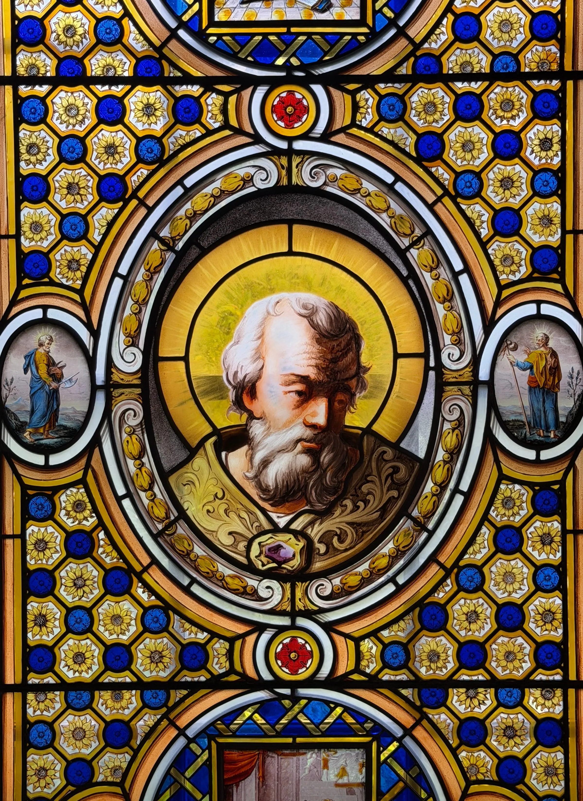 Detail of a stained glass window with a haloed bearded saint in a roundel, set in blue and gold patterned glass