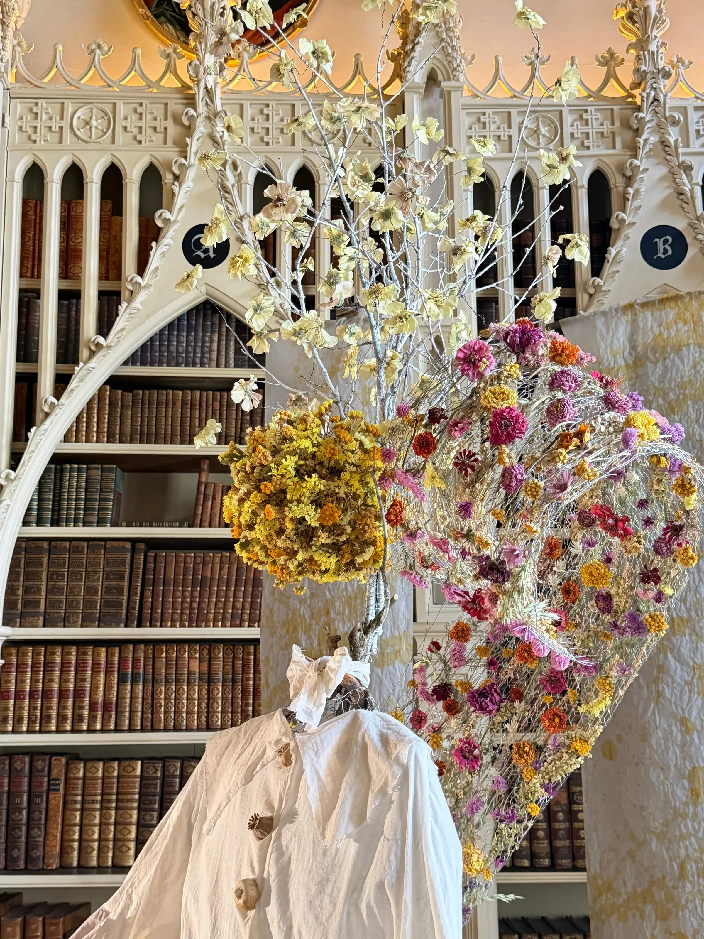 Floral installation on a dressed figure in the Library, with Gothic arched bookcases lined with leather-bound books behind