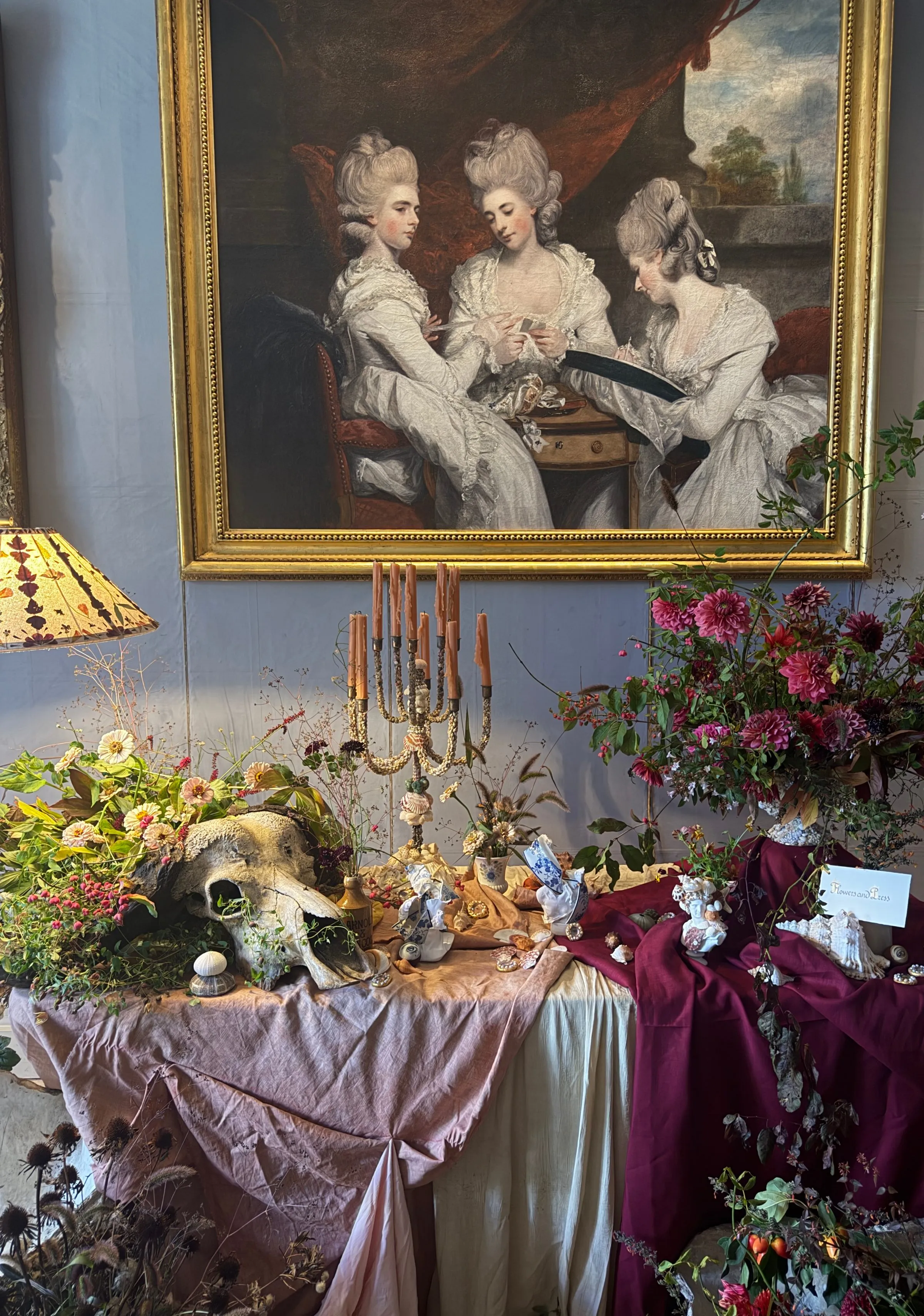 Floral table display with an animal skull and candelabra beneath an eighteenth-century portrait of three women