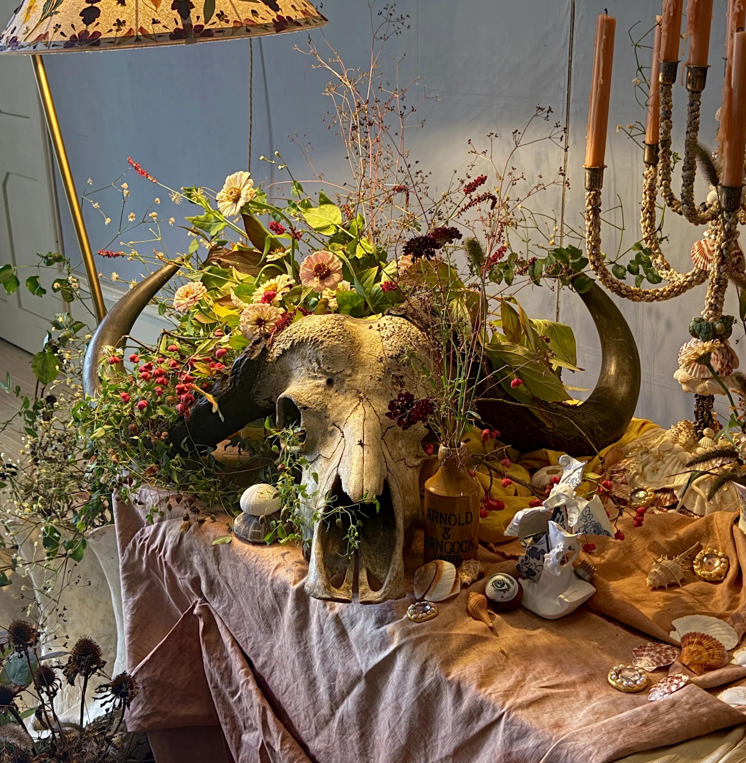 Close view of a floral arrangement spilling from a horned skull on a draped table, with gilt candelabra