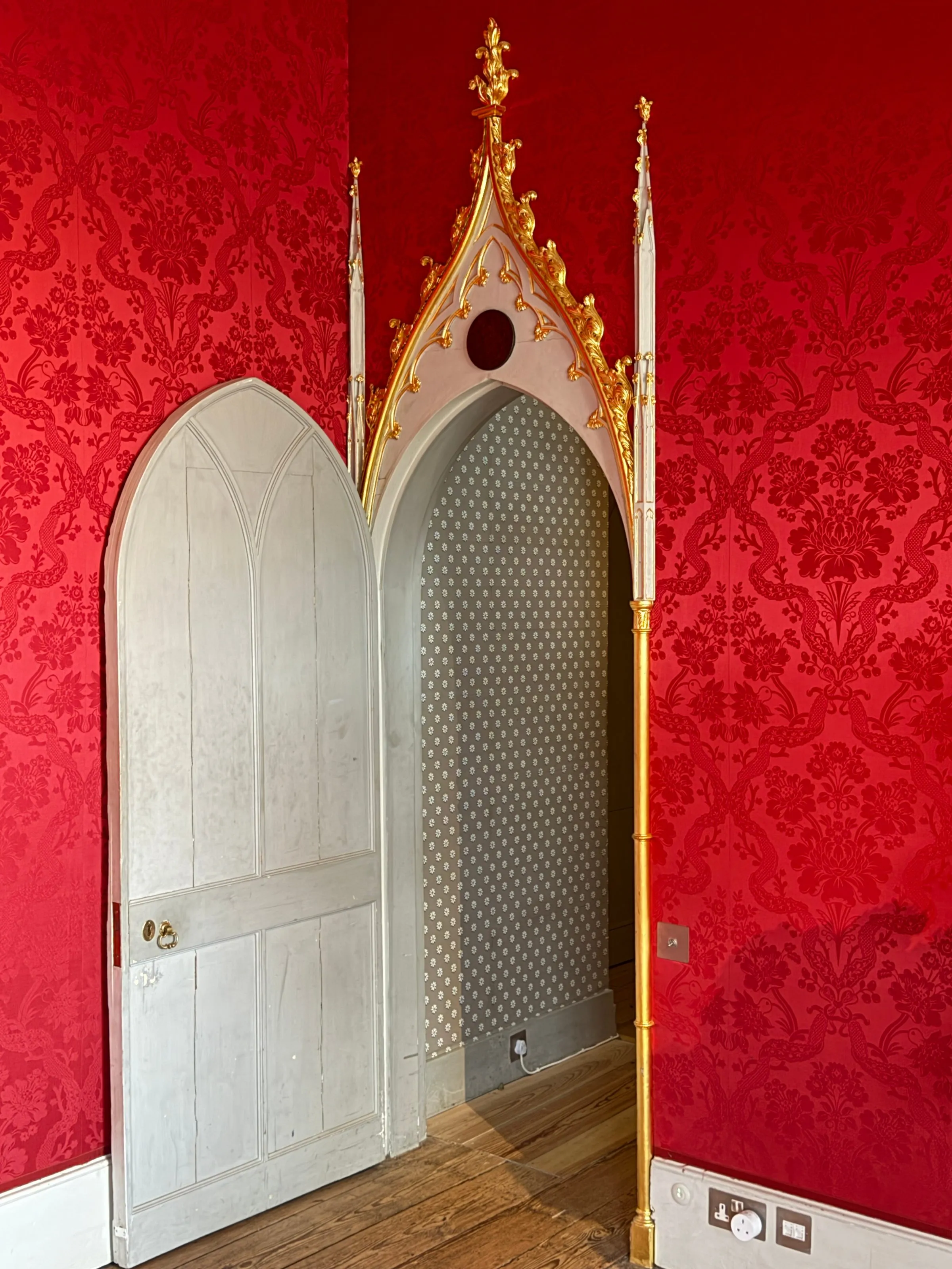 A white pointed-arch Gothic door set into crimson damask walls, crowned with a gilded pinnacle