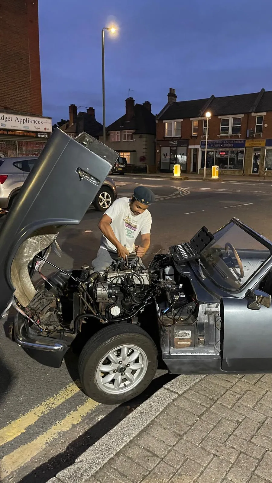 Benjamin Kändler leaning into the open engine bay of a grey Triumph Spitfire parked on a London street at dusk, the bonnet tipped forward