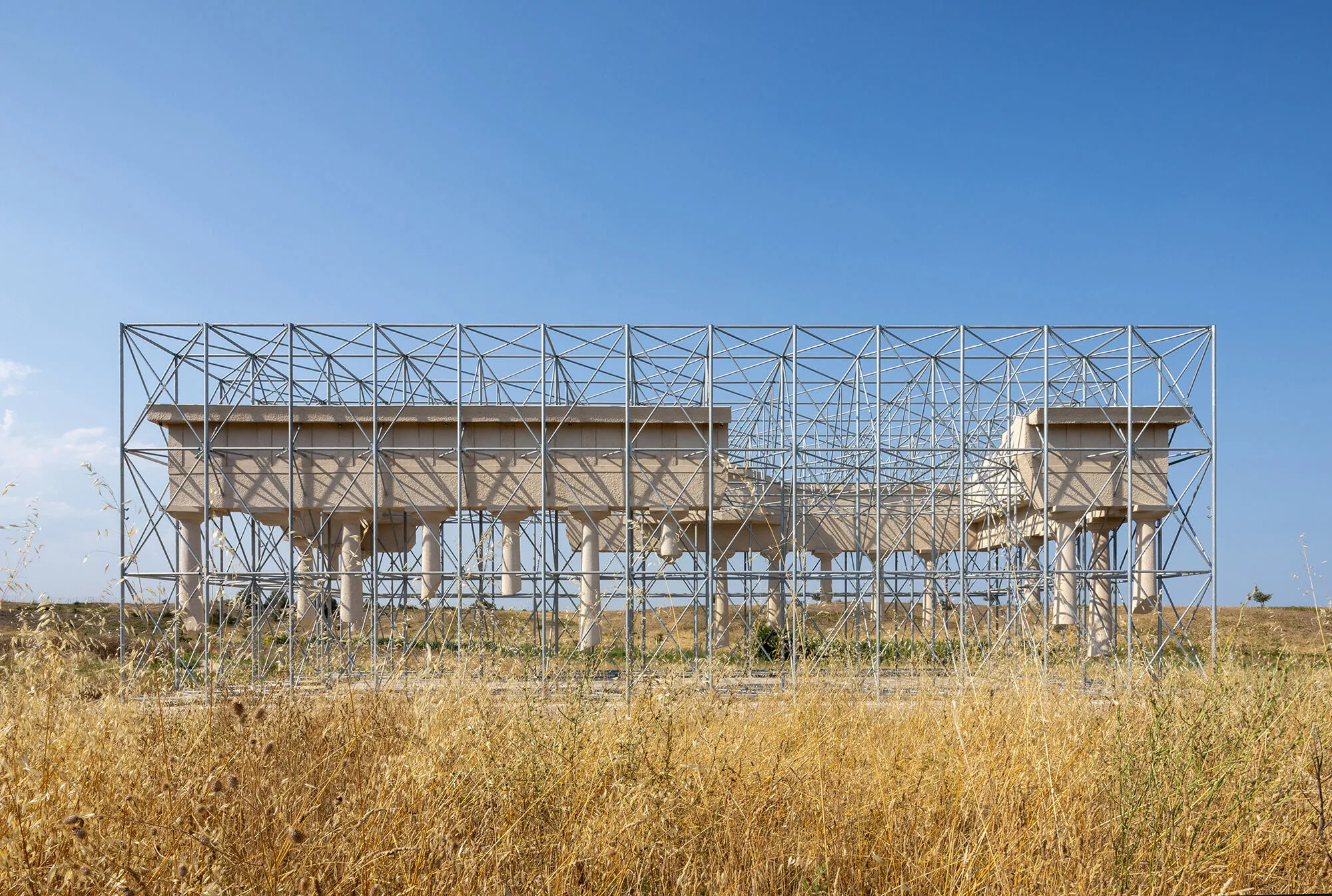 A pale stone classical temple suspended upside down inside a galvanised steel scaffold frame, standing in a field of dry golden grass under a blue sky